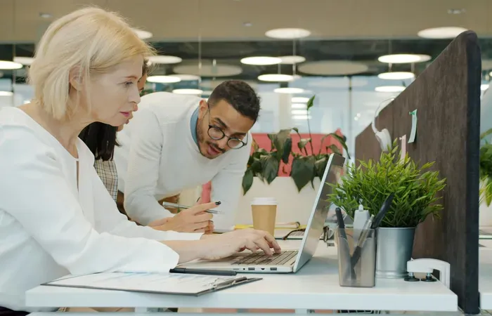 Two coworkers review a laptop at a bright modern desk with plants and coffee, highlighting service at Town Directory 360